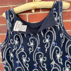 K&ouml;nnte beinhalten: Marineblaues &auml;rmelloses Kleid mit wei&szlig;en und blauen Blumenstickereien. Das Kleid ist mit Pailletten und Perlen verziert. Das Kleid h&auml;ngt an einem Vintage-Kleiderb&uuml;gel aus gelbem Stoff. Das Etikett lautet "Adrianna Papell Occasions".