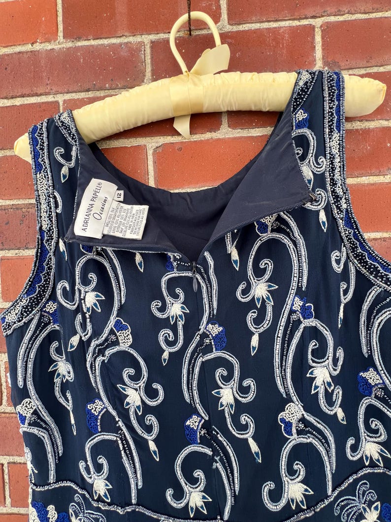 K&ouml;nnte beinhalten: Marineblaues &auml;rmelloses Kleid mit wei&szlig;en und blauen Blumenstickereien. Das Kleid ist mit Pailletten und Perlen verziert. Das Kleid h&auml;ngt an einem Vintage-Kleiderb&uuml;gel aus gelbem Stoff. Das Etikett lautet "Adrianna Papell Occasions".