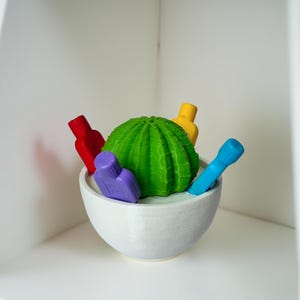 May include: A white ceramic bowl contains a green cactus-shaped ornament and several colourful, bottle-shaped items. The bottles are red, yellow, purple, and blue. The objects are arranged on a white shelf.