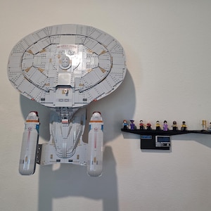 May include: A large, light gray model of a spaceship, constructed from interlocking blocks, is mounted on a wall. Below it, a black shelf displays several small figurines. The spaceship has a large, circular top section and two elongated lower sections.