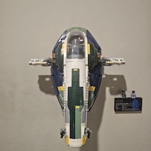 Puede incluir: Modelo LEGO detallado de una nave espacial, con ladrillos azul oscuro, verde, blanco y amarillo. El modelo está montado en una pared, con una pequeña pantalla con minifiguras y una placa.