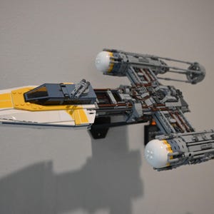 May include: A white and yellow Lego Star Wars X-wing fighter spaceship with gray details. The spaceship is mounted on a wall with a black stand.