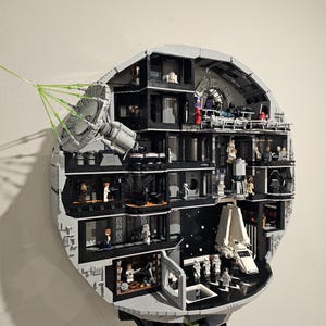 May include: A detailed LEGO model of the Death Star, showcasing interior sections with miniature figures and a white shuttle. The gray and black structure features a large dish and green laser beams extending outward. A blue plaque is visible.