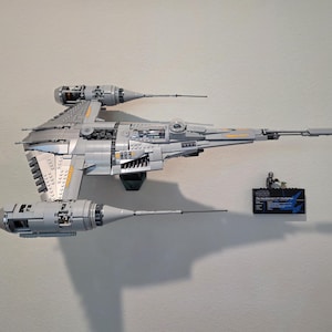 Puede incluir: Maqueta Lego gris y amarilla de una nave espacial, montada en una pared blanca. El modelo tiene dos grandes motores y una parte delantera larga y puntiaguda. Una pequeña placa negra con texto es visible debajo.
