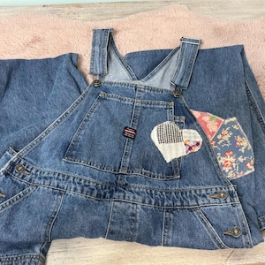 May include: Blue denim overalls with adjustable straps and multiple pockets. Features a heart-shaped patch with a black and white checkered pattern and floral accents. The overalls are made of a durable denim material.