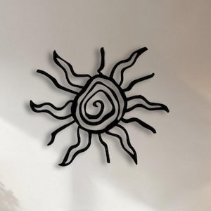 Puede incluir: Arte mural de sol de metal negro con un centro en espiral y rayos ondulados. La pieza decorativa está montada en una pared blanca sobre un sofá blanco. La habitación está decorada con tonos neutros y elementos naturales.