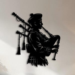 May include: Black metal wall art depicting a Scottish bagpiper in traditional attire. The silhouette shows the musician playing the bagpipes, with intricate details of the instrument and clothing. The artwork is mounted on a white wall, adding a touch of cultural flair.