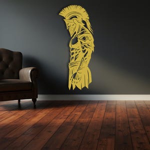 May include: A golden-colored wall art piece depicting a Spartan warrior in profile. The artwork is mounted on a dark gray wall above a wooden floor. The warrior is wearing a helmet and armor, with intricate details.
