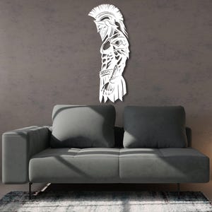 May include: White metal wall art depicting a Spartan warrior in profile. The artwork features a detailed design of the warrior's helmet, armor, and physique. The art is mounted on a gray wall above a gray sofa.