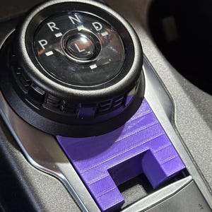 Op de afbeelding: Een paars 3D-geprint plastic afdekaccessoire op de middenconsole van een voertuig, geplaatst onder een zwarte draaiknop voor versnellingen met de letters P, R, N, D, L en boven een parkeerremknop met een P-symbool.