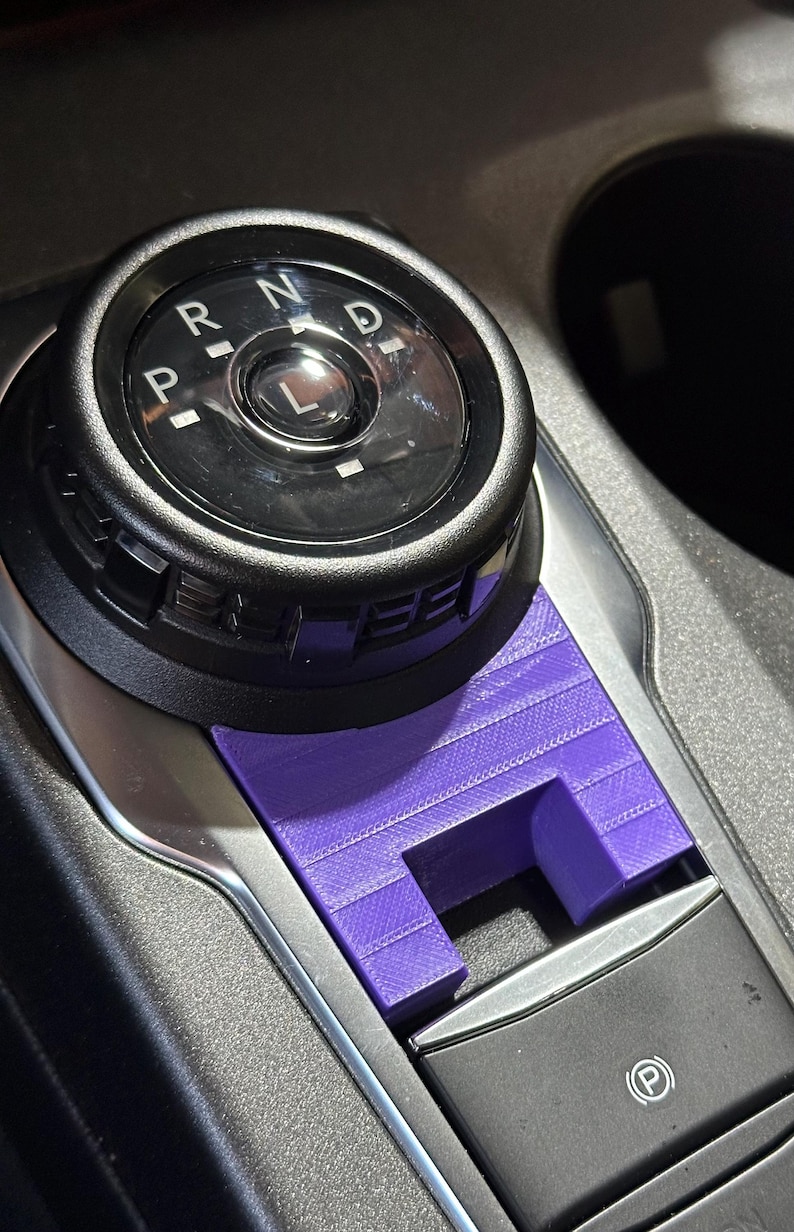 Op de afbeelding: Een paars 3D-geprint plastic afdekaccessoire op de middenconsole van een voertuig, geplaatst onder een zwarte draaiknop voor versnellingen met de letters P, R, N, D, L en boven een parkeerremknop met een P-symbool.