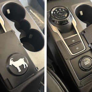 Puede incluir: Tapa de consola central negra con una silueta de cabra blanca sobre un emblema circular negro. La tapa está al lado de una palanca de cambios y portavasos. La tapa se abre para revelar botones y un dial etiquetado como "GOAT MODES".