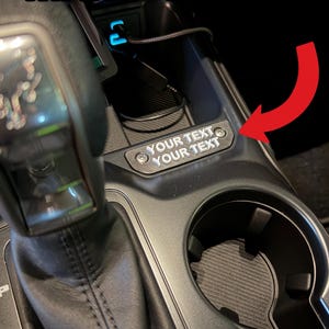 May include: Black car console with the text "MANY COLORS" in white. A red arrow points to a small, black plate with the text "YOUR TEXT YOUR TEXT". Includes a cup holder and gear shift.