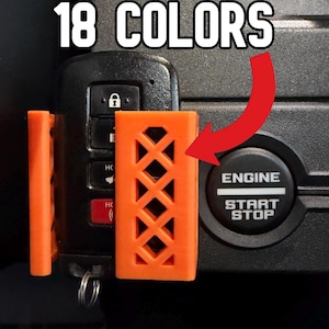 May include: An orange plastic key fob holder with a lattice pattern attached to a black vehicle key fob. The text 18 COLORS is displayed at the top, and a red arrow points from the text to the orange holder next to an engine start stop button.