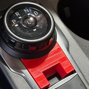 Op de afbeelding: Een felrood, 3D-geprint plastic accessoire voor de versnellingspook in een middenconsole van een voertuig. De afdekking bevindt zich onder een zwarte ronde draaiknop met de letters P, R, N, D, L en boven een knop voor de parkeerrem.