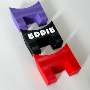 Op de afbeelding: Drie U-vormige plastic standaards in paars, zwart en rood. De zwarte standaard is voorzien van de naam EDDIE in verhoogde witte hoofdletters op de voorzijde. Elke standaard heeft een hol bovenoppervlak en een textuur van het 3D-printproces.