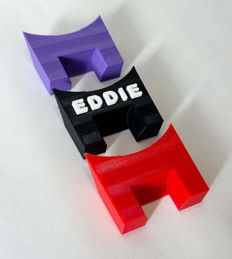 Op de afbeelding: Drie U-vormige plastic standaards in paars, zwart en rood. De zwarte standaard is voorzien van de naam EDDIE in verhoogde witte hoofdletters op de voorzijde. Elke standaard heeft een hol bovenoppervlak en een textuur van het 3D-printproces.