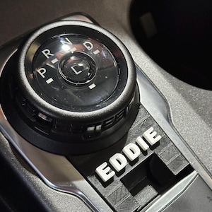 May include: A car's gear shift dial with the name "EDDIE" in white block letters. The dial is black with white markings for Park, Reverse, Neutral, Drive, and Low. The surrounding console is dark grey.