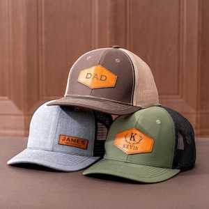 Pode incluir: Três bonés de beisebol com patches de couro personalizados. Um boné marrom tem "DAD" no patch, um boné cinza tem "JAMES GROOMSMAN" e um boné verde tem "K KEVIN" com um gráfico de chifre de veado. Cada boné tem uma parte traseira de malha.