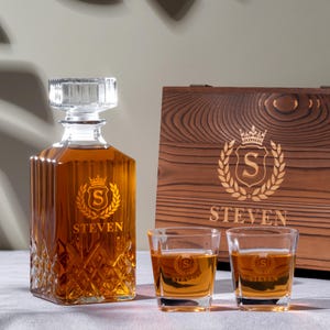 Puede incluir: Un juego de decantador de whisky personalizado con una caja de madera. El decantador y los vasos de chupito están grabados con un diseño de escudo, la letra "S" y el nombre "STEVEN". El decantador está lleno de líquido ámbar. La caja de madera tiene un diseño grabado similar.