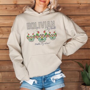 May include: A beige hoodie with a graphic print of three colorful cartoon faces and the text "Bolivian by birth, Diablo by choice!"