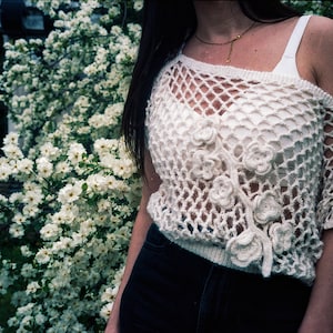 Peut inclure: Un haut en crochet blanc avec un motif floral délicat et une maille ajourée. Le haut est ample et légèrement décolleté.