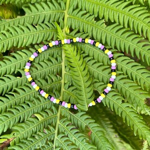 May include: A beaded bracelet with yellow, black, white, and purple beads. The bracelet is on a green fern leaf.