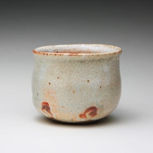 Peut inclure: Une petite tasse en céramique arrondie avec un émail blanc cassé texturé présentant de subtils motifs de craquelures. La surface extérieure affiche des taches brunes éparses et des taches brun-rouge distinctes près de la base.