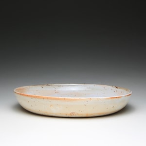 Peut inclure: Un bol en céramique rond et peu profond avec un émail blanc cassé et crème moucheté. Le bord présente des tons orange-brun subtils et la surface est parsemée de petites taches brun foncé. Une petite marque de potier circulaire est visible.