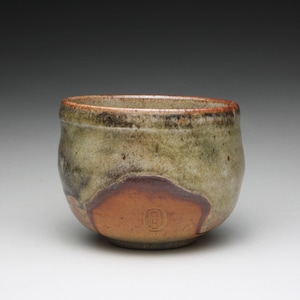 May include: A ceramic bowl with a rounded shape, featuring a mottled glaze in shades of olive green, brown, and grey. The lower portion displays a smooth, unglazed terracotta-colored patch with a small, stamped oval maker's mark in the center.