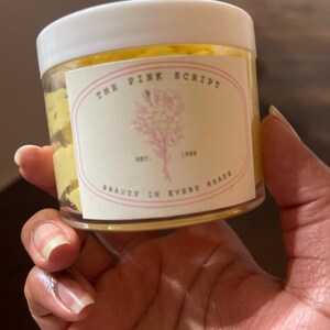 May include: A clear jar with a white lid, filled with a yellow substance. The jar has a label with the text "THE PINK SCRIPT" and a pink floral design. The label also includes the text "EST. 1996 BEAUTY IN EVERY SHADE."