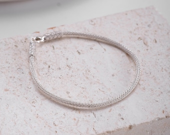 Könnte beinhalten: Ein silbernes Kettenarmband mit einem zarten, gewebten Design. Das Armband liegt auf einer weißen, strukturierten Oberfläche.
