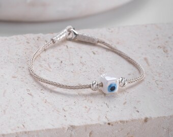 May include: A silver bracelet with a white evil eye charm. The bracelet is made of a braided silver chain and has a lobster clasp.