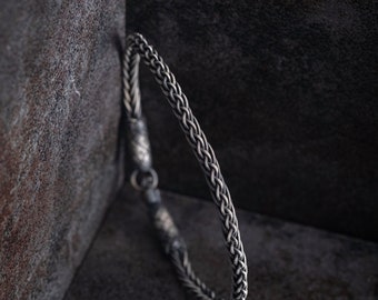 Könnte beinhalten: Ein silbernes Kettenarmband mit geflochtenem Design. Das Armband liegt auf einer dunkelgrauen, strukturierten Oberfläche.