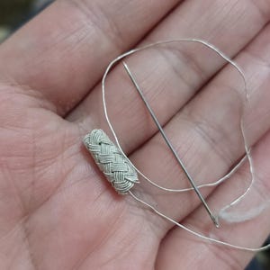 May include: A close-up of a silver braided cord with a needle and thread. The cord is woven in a tight, intricate pattern.