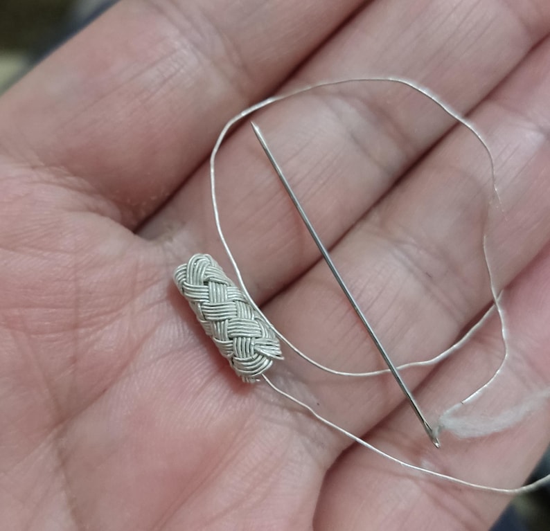 May include: A close-up of a silver braided cord with a needle and thread. The cord is woven in a tight, intricate pattern.