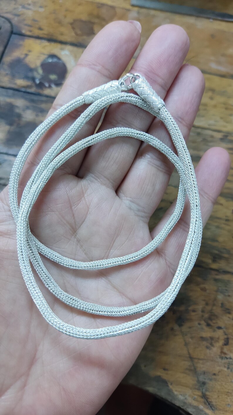 May include: A silver chain necklace with a braided design. The chain is coiled and laid out on a hand.