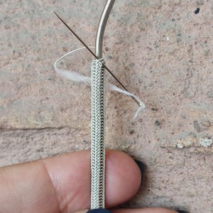 May include: A close-up of a silver chain being threaded with white thread using a needle and a metal tool.