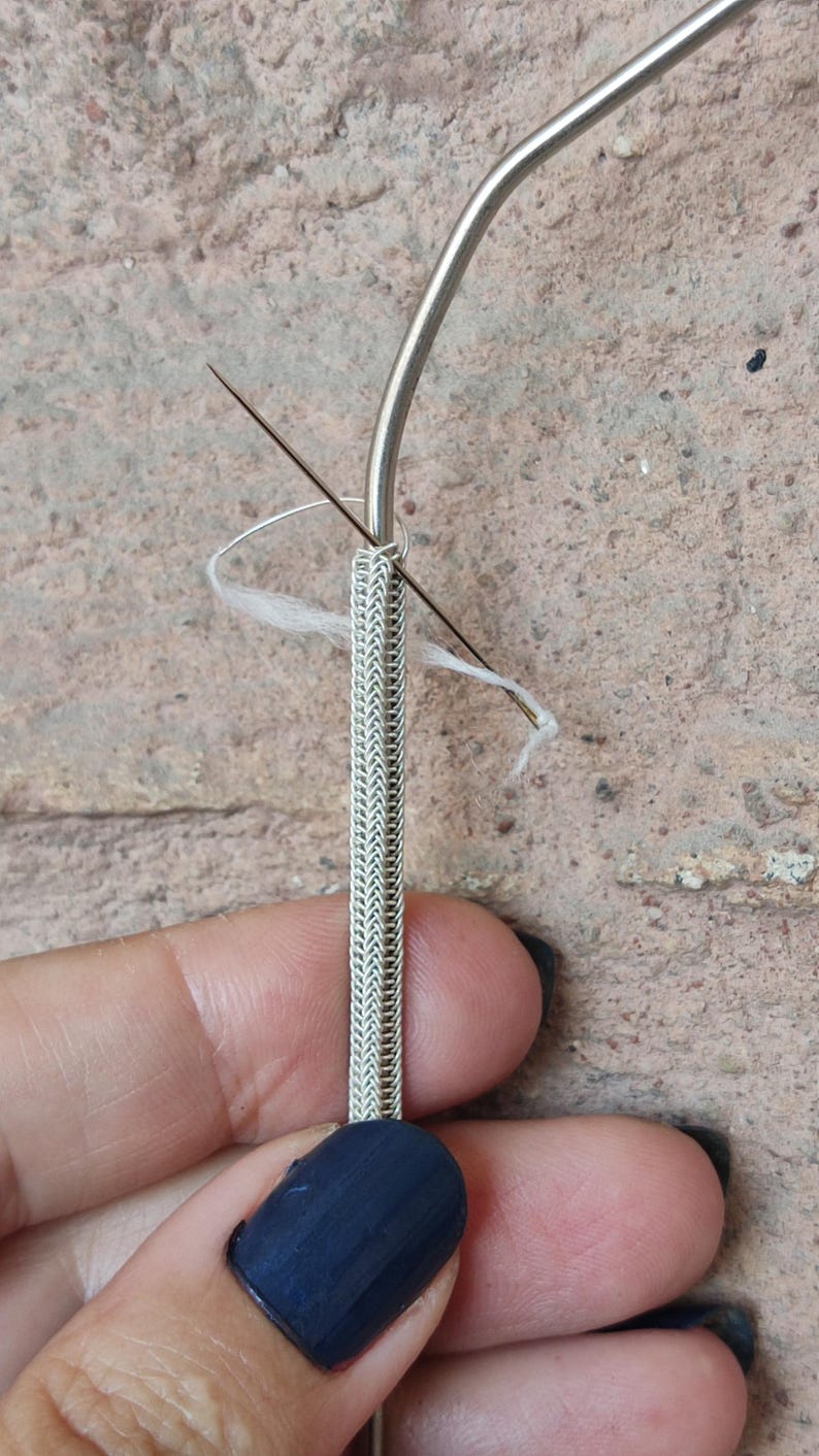May include: A close-up of a silver chain being threaded with white thread using a needle and a metal tool.