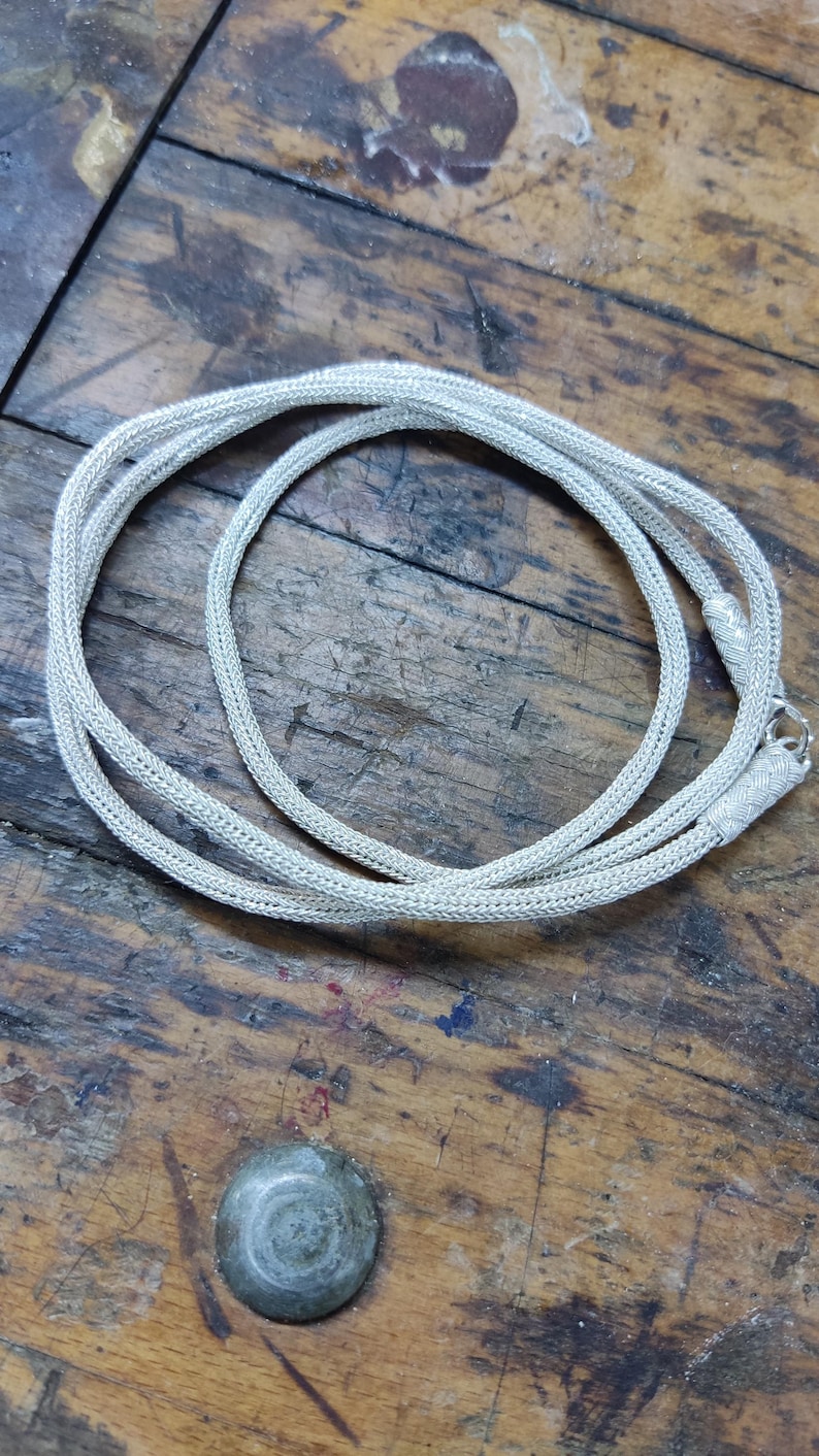 May include: A silver chain necklace with a braided design. The chain is lying on a wooden surface.