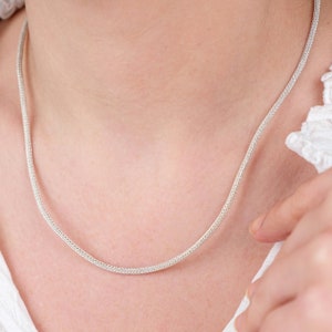 K&ouml;nnte beinhalten: Eine silberne Kettenkette mit einem zarten, gewebten Design. Die Kette ist d&uuml;nn und liegt flach auf der Haut.