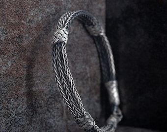 Op de afbeelding: Een zilveren gevlochten armband met een geweven ontwerp. De armband is gemaakt van meerdere zilveren draden die met elkaar zijn gevlochten om een uniek en ingewikkeld patroon te creëren.