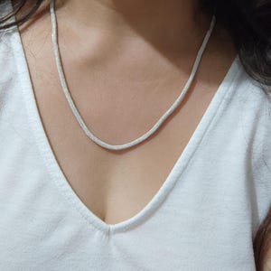 May include: A silver chain necklace with a delicate, woven design. The necklace is worn by a person with a white shirt.