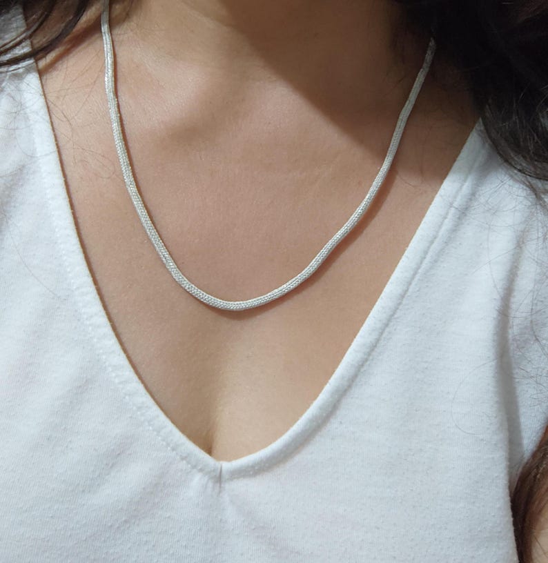 May include: A silver chain necklace with a delicate, woven design. The necklace is worn by a person with a white shirt.
