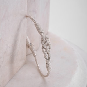 May include: A silver bracelet with a braided knot design. The bracelet is on a white marble surface.