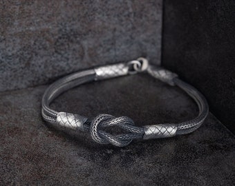 May include: A silver bracelet with a knot design. The bracelet features a woven pattern and a secure clasp. The knot is the focal point, adding a unique touch to the jewelry. The bracelet is displayed on a dark, textured surface.