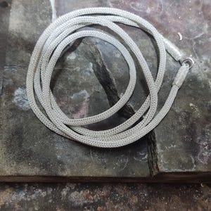 May include: A silver chain necklace with a woven design. The chain is coiled on a dark gray surface.