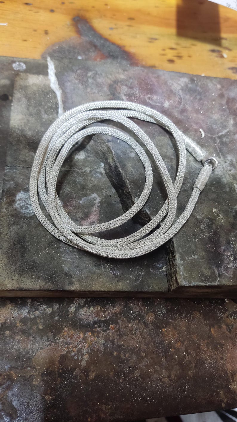 May include: A silver chain necklace with a woven design. The chain is coiled on a dark gray surface.