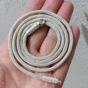 May include: A silver chain necklace with a woven design. The chain is coiled in a hand.