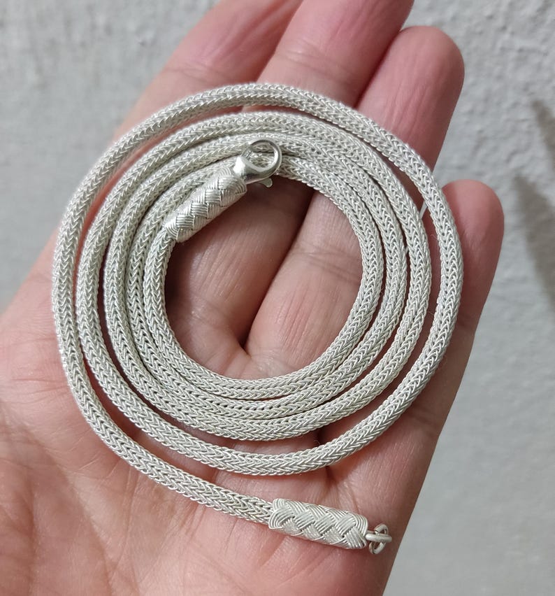 May include: A silver chain necklace with a woven design. The chain is coiled in a hand.
